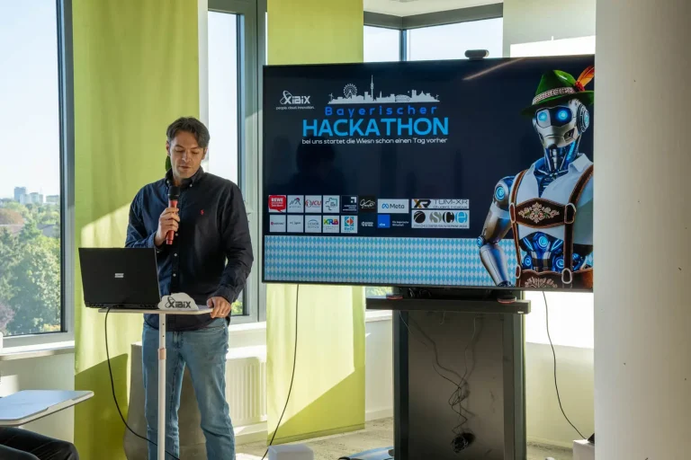 Sprecher am XIBIX Hackathon spricht in sein Mikrofon, im Hintergrund ist ein Bildschirm mit dem Titelbild der Veranstaltung