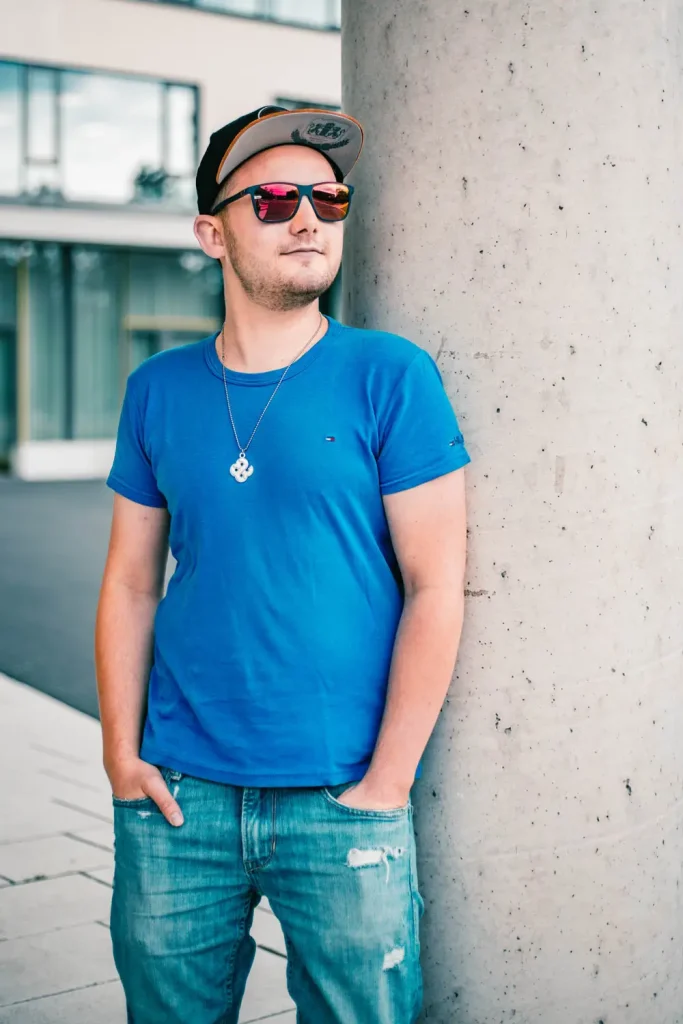 Foto eines Mannes in blauem Shirt, Sonnenbrille und Cap an einer Säule lehnend