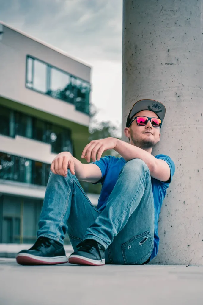 Foto eines Mannes in blauem Shirt, Sonnenbrille und Cap an einer Säule lehnend, Frontalansicht