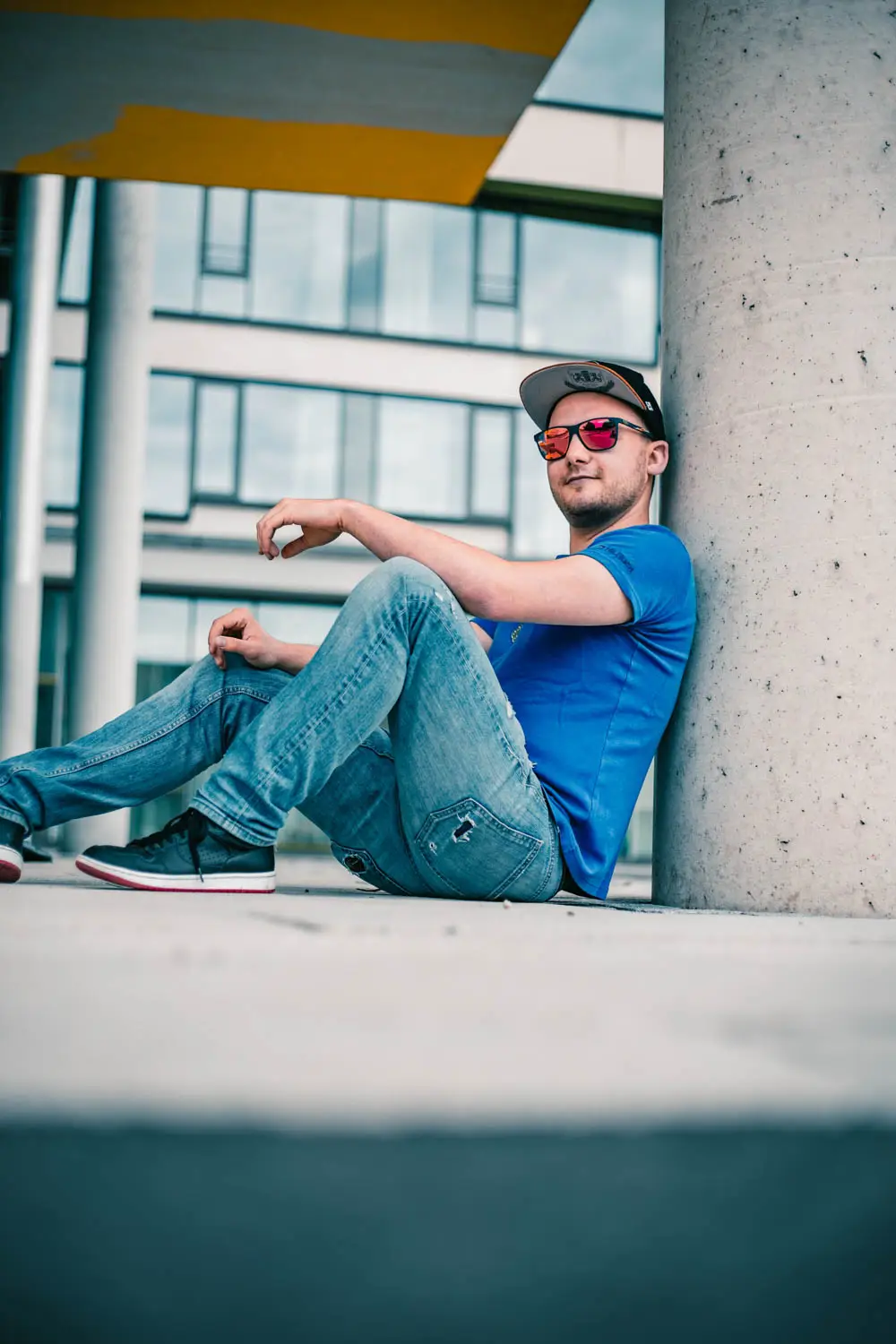 Foto eines Mannes in blauem Shirt, Sonnenbrille und Cap an einer Säule lehnend
