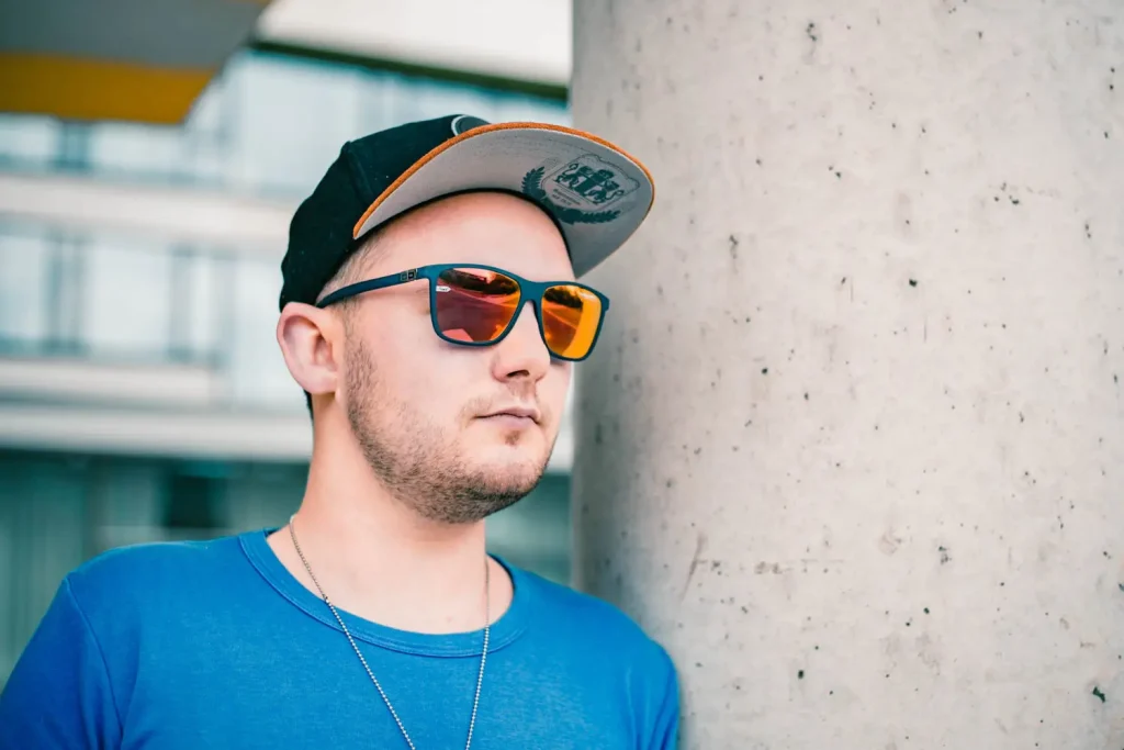 Foto eines Mannes in blauem Shirt, Sonnenbrille und Cap an einer Säule lehnend
