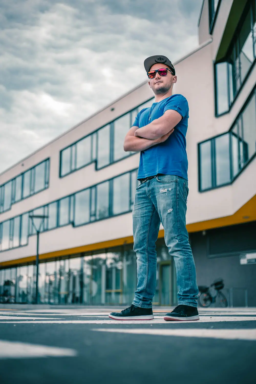 Foto eines Mannes in blauem Shirt, Sonnenbrille und Cap stehend, im Hintergrund ein Gebäudekomplex